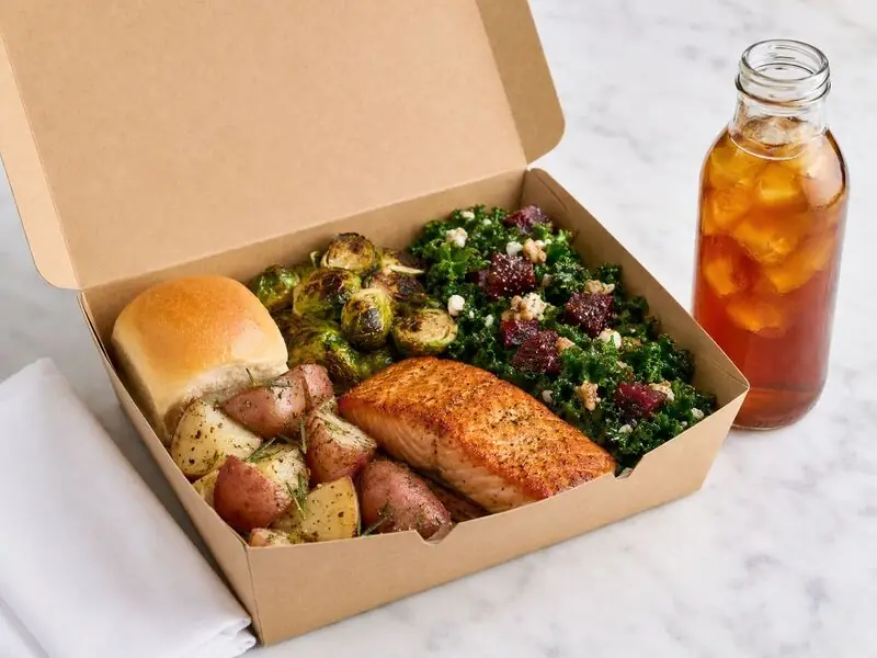Pleasanton individually packaged meal featuring grilled salmon, herb-roasted red potatoes, greens, vegetables, bread, and a chilled beverage.