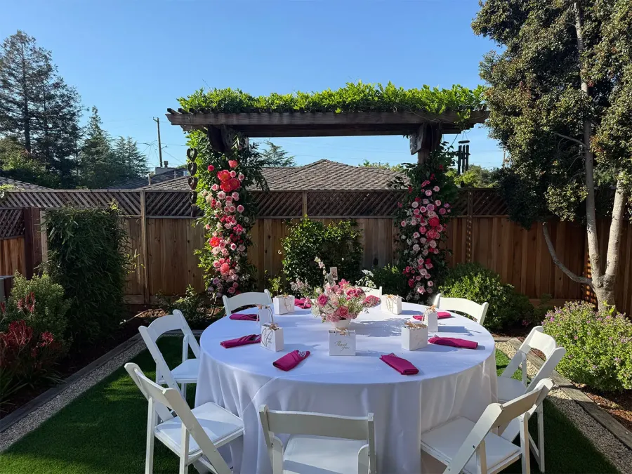 Rose arch in blush and deep red arching over a round garden table with white linen and fuchsia napkins — floral arch design for Pleasanton catering weddings
